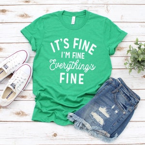 May include: Green t-shirt with white text that reads "It's Fine I'm Fine Everything's Fine". The shirt is paired with denim shorts and white sneakers.