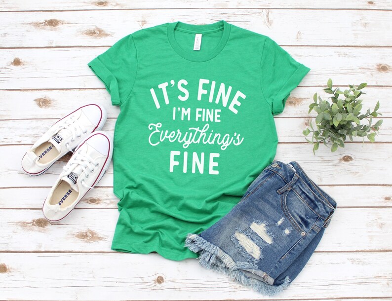 May include: Green t-shirt with white text that reads "It's Fine I'm Fine Everything's Fine". The shirt is paired with denim shorts and white sneakers.
