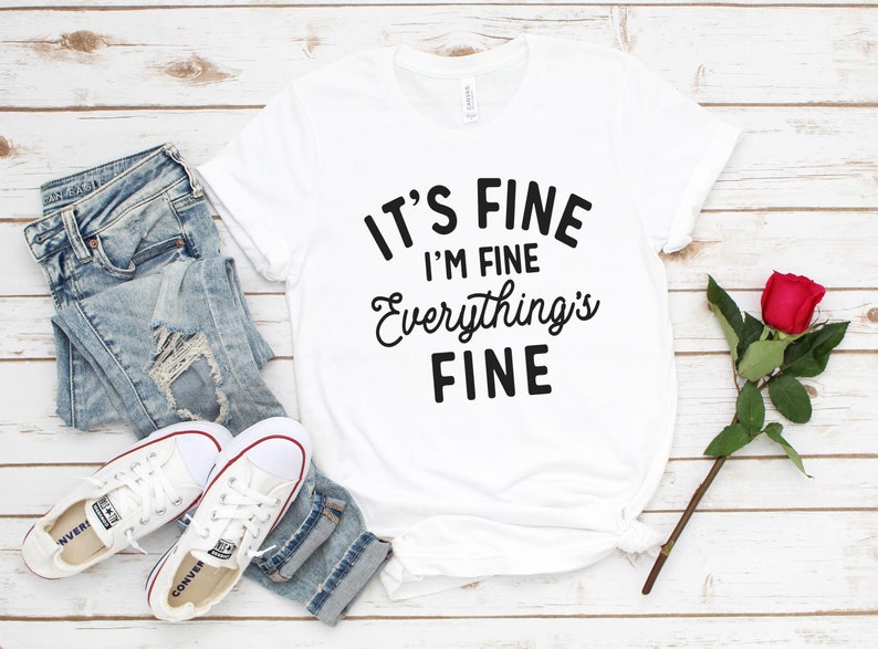 May include: White t-shirt with black text that reads "It's Fine I'm Fine Everything's Fine". The shirt is laying on a white wooden surface with a pair of white sneakers, blue jeans, and a red rose.