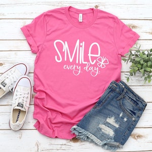 May include: Pink t-shirt with white text that says "Smile every day." The shirt is laying on a white wooden surface with a pair of white sneakers and a pair of blue denim shorts.
