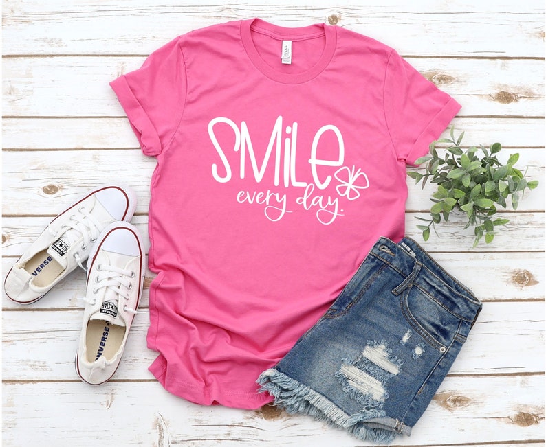May include: Pink t-shirt with white text that says "Smile every day." The shirt is laying on a white wooden surface with a pair of white sneakers and a pair of blue denim shorts.