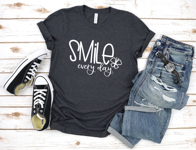 May include: A dark heather grey t-shirt with the text "SMILE every day" in white lettering and a small white four-leaf clover graphic.