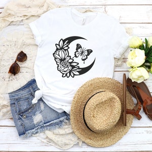 May include: White t-shirt featuring a black crescent moon design with flowers and a butterfly. The image also includes denim shorts, a straw hat, sunglasses, and brown sandals. The items are arranged on a white wooden surface.