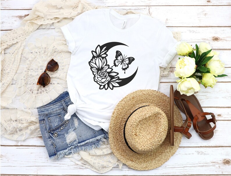 May include: White t-shirt featuring a black crescent moon design with flowers and a butterfly. The image also includes denim shorts, a straw hat, sunglasses, and brown sandals. The items are arranged on a white wooden surface.