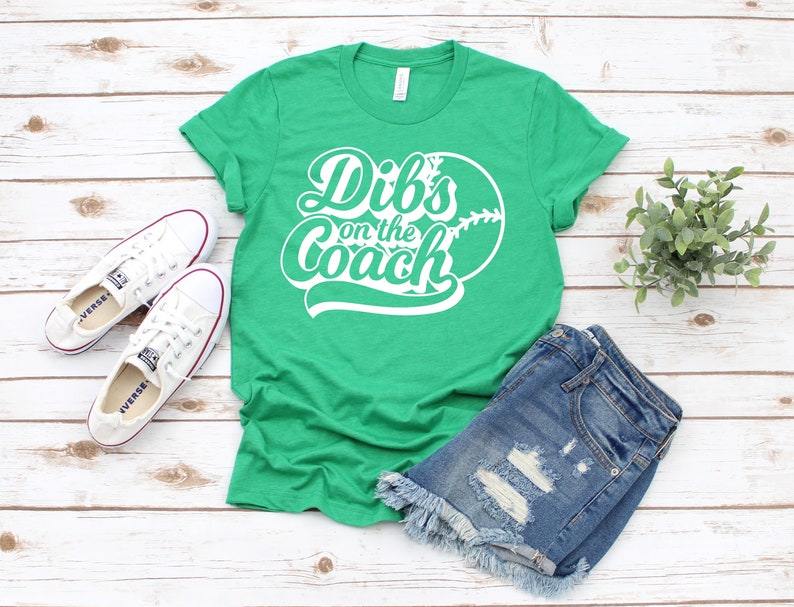 May include: Green t-shirt with white text that says "Dibs on the Coach" and a white baseball graphic.