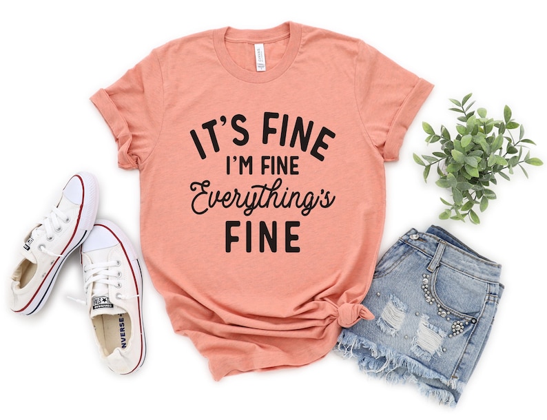 May include: A peach colored t-shirt with black text that reads "It's Fine I'm Fine Everything's Fine".