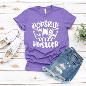 May include: Purple t-shirt with white text that reads "Popsicle Hustler" and a graphic of popsicles.