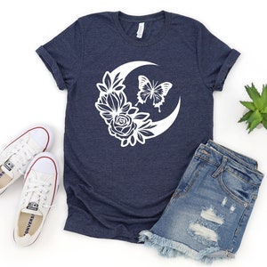 May include: Navy blue t-shirt featuring a white crescent moon design with floral and butterfly accents. The shirt is paired with denim shorts and white canvas sneakers. A small succulent plant is also visible.