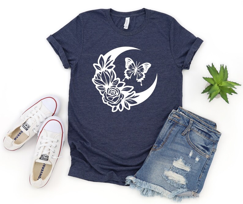 May include: Navy blue t-shirt featuring a white crescent moon design with floral and butterfly accents. The shirt is paired with denim shorts and white canvas sneakers. A small succulent plant is also visible.