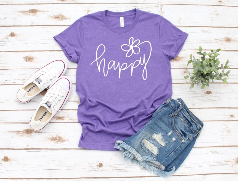May include: Purple t-shirt with a white graphic that says "happy" with a flower design. The shirt is laying on a white wooden surface with a pair of white sneakers and denim shorts.