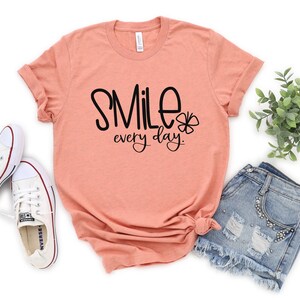 May include: A peach colored t-shirt with the text "Smile every day." in black lettering and a small four-leaf clover graphic.