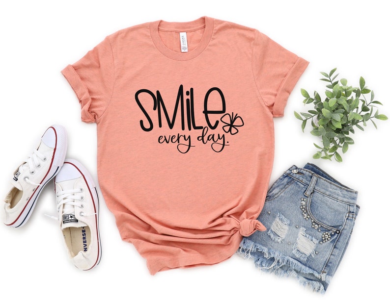 May include: A peach colored t-shirt with the text "Smile every day." in black lettering and a small four-leaf clover graphic.
