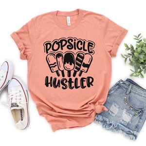 May include: A peach colored t-shirt with black text that reads "Popsicle Hustler" and a graphic of four popsicles.