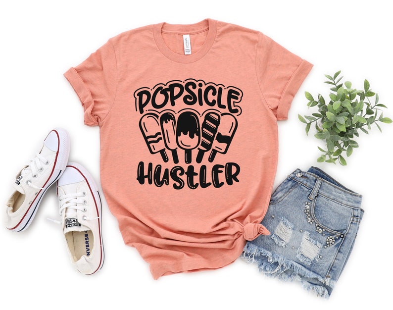 May include: A peach colored t-shirt with black text that reads "Popsicle Hustler" and a graphic of four popsicles.