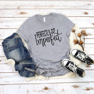Puede incluir: Una camiseta gris con el texto "Perfectly Imperfect" en letras negras con un peque&ntilde;o s&iacute;mbolo de coraz&oacute;n. La camiseta est&aacute; colocada sobre una superficie de madera blanca con un par de pantalones vaqueros azules y zapatillas negras.