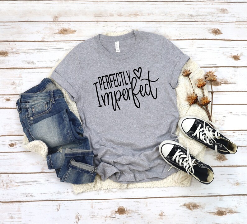 Puede incluir: Una camiseta gris con el texto "Perfectly Imperfect" en letras negras con un peque&ntilde;o s&iacute;mbolo de coraz&oacute;n. La camiseta est&aacute; colocada sobre una superficie de madera blanca con un par de pantalones vaqueros azules y zapatillas negras.