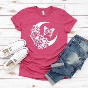 May include: A maroon t-shirt featuring a white crescent moon design with a butterfly and floral elements. The shirt is paired with blue jeans and white sneakers. A casual, everyday outfit.