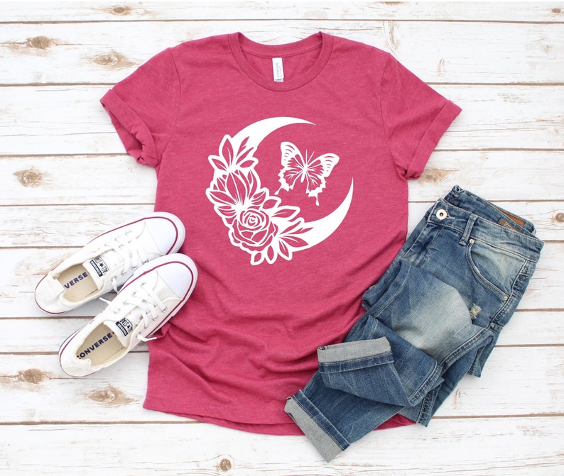 May include: A maroon t-shirt featuring a white crescent moon design with a butterfly and floral elements. The shirt is paired with blue jeans and white sneakers. A casual, everyday outfit.