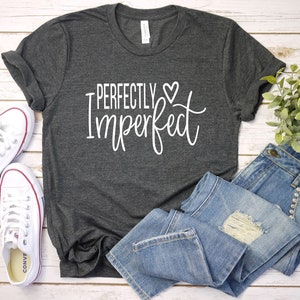 Puede incluir: Una camiseta gris oscuro con el texto "Perfectly Imperfect" en letras blancas con un peque&ntilde;o s&iacute;mbolo de coraz&oacute;n. La camiseta est&aacute; colocada sobre una superficie de madera blanca con un par de zapatillas blancas y unos vaqueros azules.
