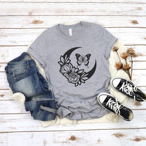 May include: A gray t-shirt featuring a black graphic of a crescent moon, flowers, and a butterfly. The shirt is paired with blue jeans and black canvas sneakers with white soles and laces. The items are arranged on a white wooden surface.