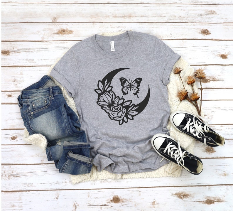 May include: A gray t-shirt featuring a black graphic of a crescent moon, flowers, and a butterfly. The shirt is paired with blue jeans and black canvas sneakers with white soles and laces. The items are arranged on a white wooden surface.
