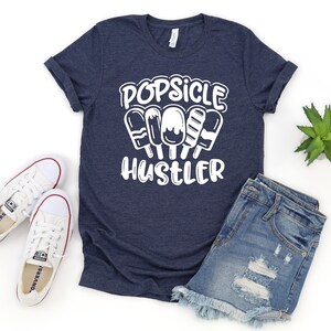 May include: A navy blue t-shirt with white text that reads "Popsicle Hustler". The text is surrounded by a graphic of four popsicles.