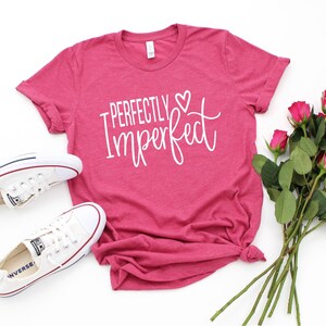 Puede incluir: Una camiseta rosa con el texto "Perfectly Imperfect" en letras blancas con un peque&ntilde;o s&iacute;mbolo de coraz&oacute;n. La camiseta est&aacute; colocada sobre una superficie blanca con un par de zapatillas blancas y un ramo de rosas rosas.