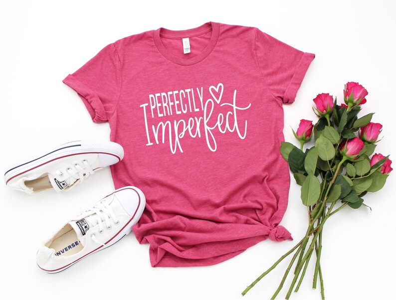 Puede incluir: Una camiseta rosa con el texto "Perfectly Imperfect" en letras blancas con un peque&ntilde;o s&iacute;mbolo de coraz&oacute;n. La camiseta est&aacute; colocada sobre una superficie blanca con un par de zapatillas blancas y un ramo de rosas rosas.