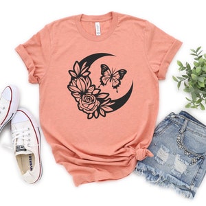 May include: Peach-colored t-shirt featuring a black crescent moon design with a butterfly and floral accents. The shirt is paired with denim shorts and white sneakers, creating a casual, everyday look. The design is centered on the shirt.