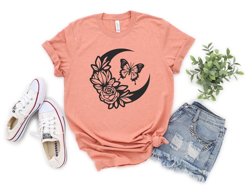 May include: Peach-colored t-shirt featuring a black crescent moon design with a butterfly and floral accents. The shirt is paired with denim shorts and white sneakers, creating a casual, everyday look. The design is centered on the shirt.