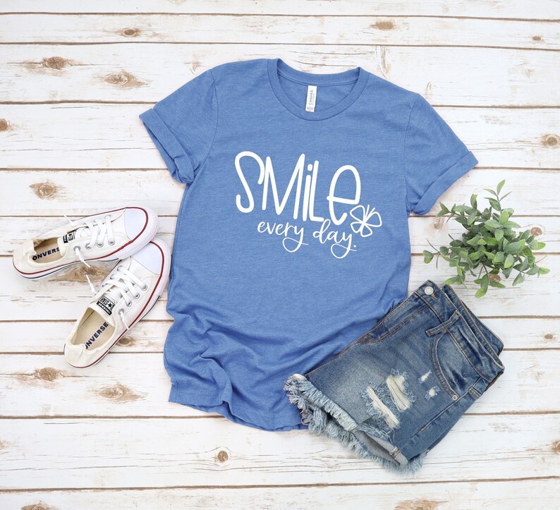 May include: A blue t-shirt with the text "Smile every day" in white lettering and a small flower graphic. The shirt is laid out on a white wood background with a pair of white sneakers and denim shorts.