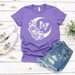May include: A purple t-shirt with a white floral and butterfly design, paired with denim shorts and white sneakers. The shirt features a crescent moon shape with flowers and a butterfly. The outfit is laid out on a wooden surface.