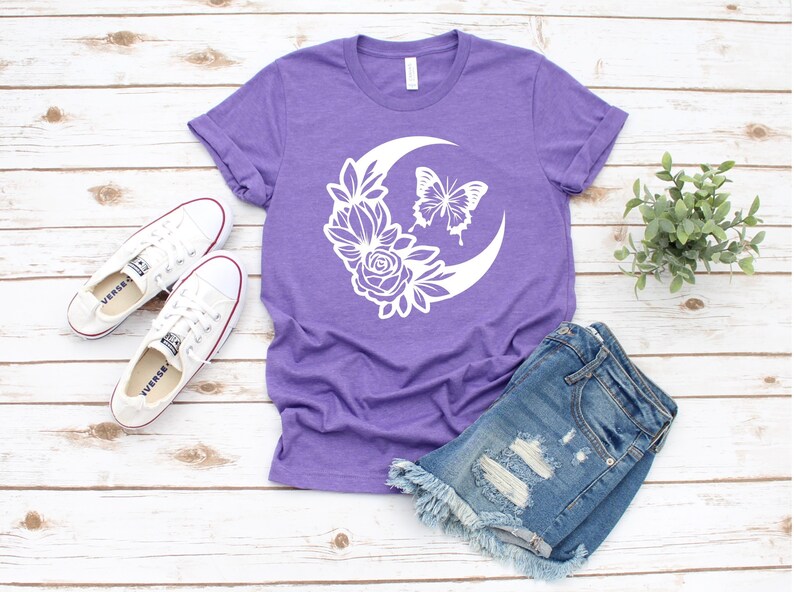 May include: A purple t-shirt with a white floral and butterfly design, paired with denim shorts and white sneakers. The shirt features a crescent moon shape with flowers and a butterfly. The outfit is laid out on a wooden surface.