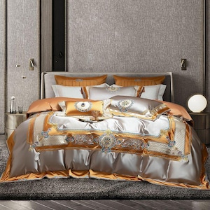 May include: A luxurious bed set featuring a silver comforter with gold and blue embroidered details. The set includes matching pillowcases and decorative pillows in gold and silver tones, creating an elegant bedroom aesthetic.