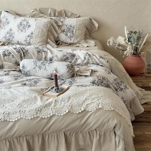 May include: A beige floral bedding set with a ruffled edge. The set includes two square pillows with lace trim, a bolster pillow, and a duvet cover. A lit candle sits on a small wooden tray. A vase with dried flowers is on the right.