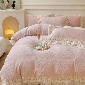 Pode incluir: Um conjunto de roupa de cama rosa com padr&atilde;o floral e acabamento em renda. O conjunto inclui uma capa de edred&atilde;o e v&aacute;rias almofadas. Uma ta&ccedil;a de champanhe e uma pequena bandeja com objetos decorativos est&atilde;o na cama.