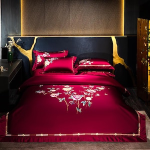Peut inclure: Une parure de lit bordeaux luxueuse avec des motifs floraux brodés. L'ensemble comprend une housse de couette et plusieurs taies d'oreiller. La literie a une bordure dorée et est présentée sur un lit avec une tête de lit sombre.