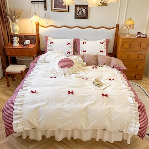May include: A white and red gingham patterned bedding set with ruffled edges. The set includes a duvet cover, pillowcases, and decorative pillows. Red bows are scattered across the duvet. The bed is set in a room with wooden furniture.