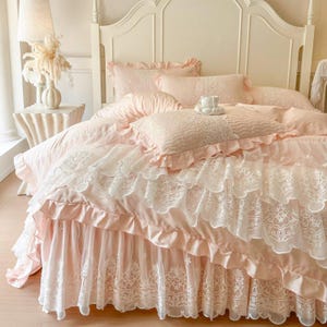 May include: A bed with a pink and white lace bedding set. The set includes a ruffled duvet cover, pillows, and a bed skirt. The headboard is white, and the overall aesthetic is romantic and feminine.