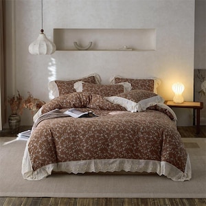 Op de afbeelding: Een bruine bloemen beddengoed set met witte kanten rand. De set bevat een dekbedovertrek, kussenslopen en sierkussens. Het beddengoed ligt op een bed met een neutraal tapijt en een lamp.