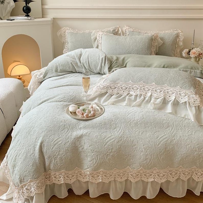 Pode incluir: Um conjunto de colcha e almofadas verde claro com um padr&atilde;o floral e acabamento em renda. A roupa de cama inclui uma capa de edred&atilde;o, almofadas e uma saia de cama. Uma bandeja de prata com guloseimas e uma ta&ccedil;a de champanhe est&atilde;o na cama.