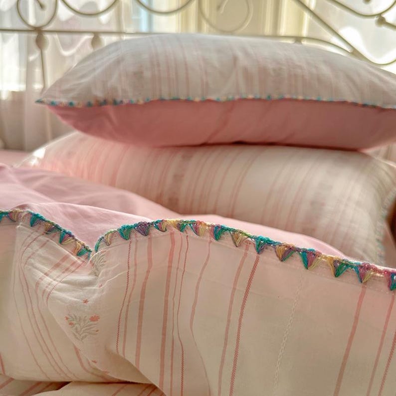 May include: A bed with pink and white striped bedding. The pillowcases and duvet cover feature a decorative trim with colourful stitching. The pillows are stacked. The bedding is in a bedroom.