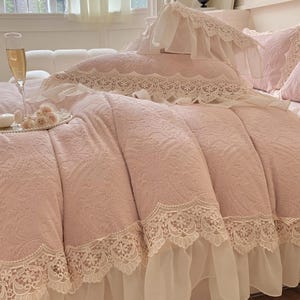 Pode incluir: Um edredom rosa com um padr&atilde;o floral e acabamento em renda branca. A cama &eacute; feita com travesseiros combinando e uma fronha decorativa. Uma ta&ccedil;a de champanhe e um prato pequeno com flores est&atilde;o na cama.