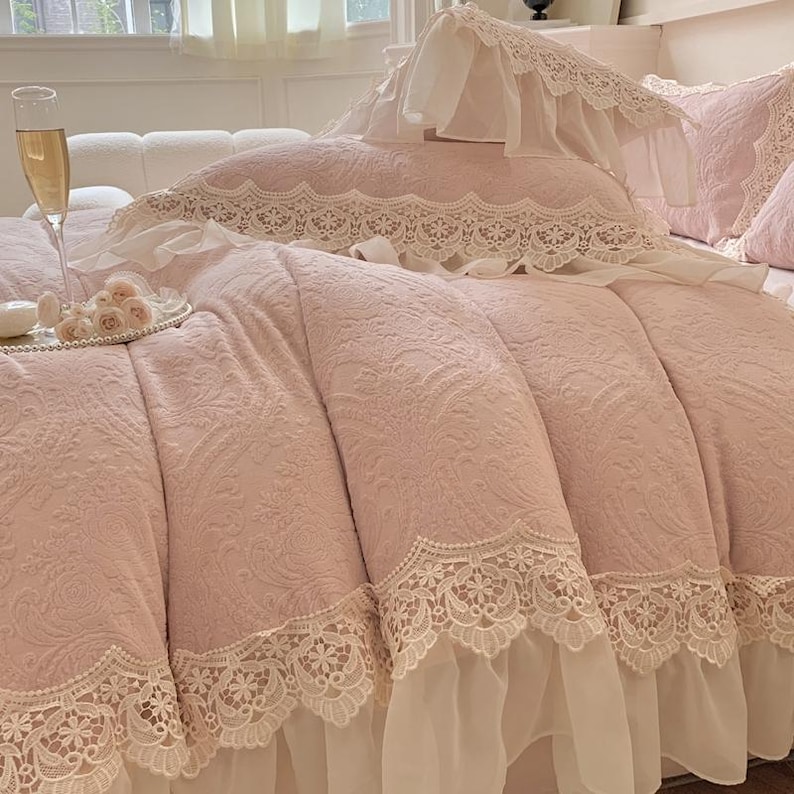 Pode incluir: Um edredom rosa com um padr&atilde;o floral e acabamento em renda branca. A cama &eacute; feita com travesseiros combinando e uma fronha decorativa. Uma ta&ccedil;a de champanhe e um prato pequeno com flores est&atilde;o na cama.