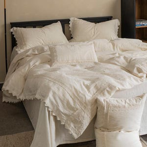 May include: A cream-coloured bed set featuring a duvet cover and pillowcases with a scalloped edge. The bedding includes several pillows of different sizes, creating a layered and inviting appearance. The dark bed frame contrasts with the light-coloured linens.