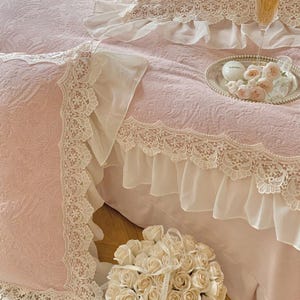 Pode incluir: Conjunto de roupa de cama rosa claro com detalhes delicados em renda e bordas com babados. O conjunto inclui uma fronha e uma saia de cama. Um buqu&ecirc; de rosas brancas est&aacute; em primeiro plano. Uma bandeja de prata com rosas e uma ta&ccedil;a de champanhe est&aacute; na cama.