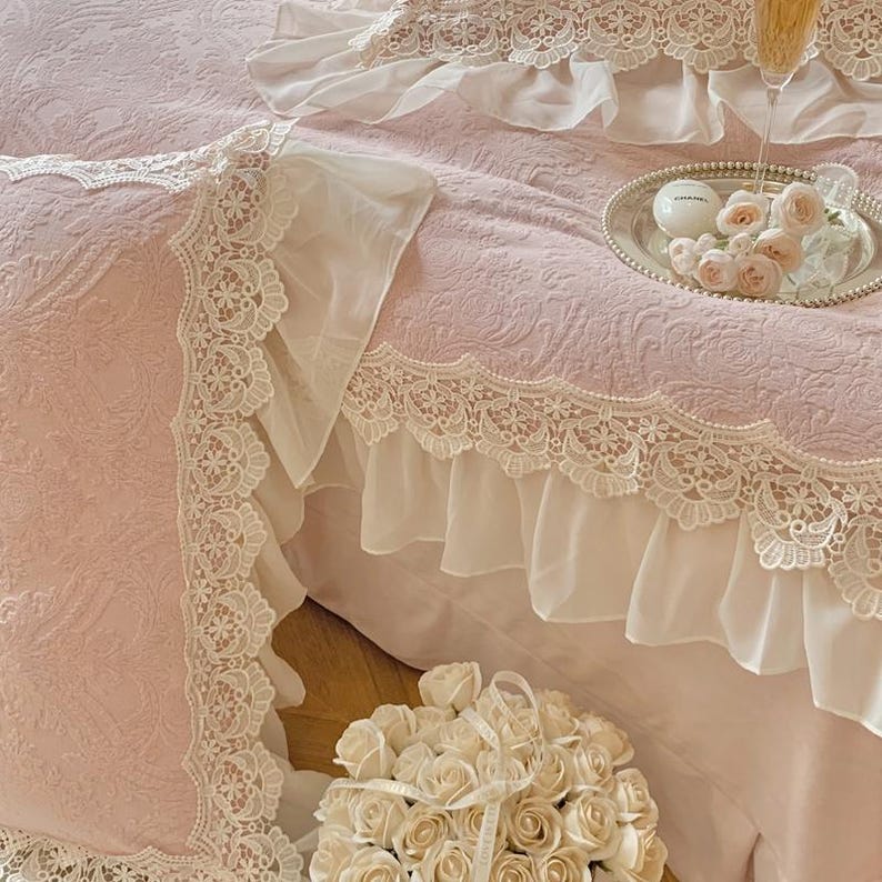 Pode incluir: Conjunto de roupa de cama rosa claro com detalhes delicados em renda e bordas com babados. O conjunto inclui uma fronha e uma saia de cama. Um buqu&ecirc; de rosas brancas est&aacute; em primeiro plano. Uma bandeja de prata com rosas e uma ta&ccedil;a de champanhe est&aacute; na cama.