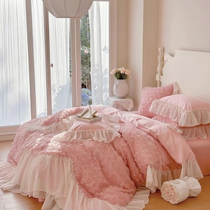 May include: A pink bed set with a floral pattern and ruffled trim. The bedding includes a comforter, pillows, and a bed skirt. The room has a window with sheer curtains and a white vase with flowers.