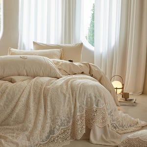 Puede incluir: Un juego de cama beige con una superposición de encaje. La ropa de cama incluye almohadas, un edredón y una colcha. La cama está frente a una ventana con cortinas blancas. Se ve una lámpara pequeña y una mesita.