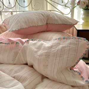 May include: Close-up of a bed featuring a white and pink striped duvet cover and pillowcases. The pillowcases have a pink border and decorative trim. The bedding has a floral pattern and is arranged on a bed frame.